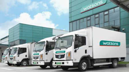 AS Watson boosts sustainability with EV fleet and waste reduction plans White delivery trucks with green Watsons logo on the side, in front of building with blue sky and white clouds in background