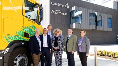 DHL switches to HVO for sustainable transportation of pharmaceutical goods DHL Global Forwarding Freight Ireland and Alexion AstraZeneca Rare Disease standing in front of Alexion AstraZeneca Rare Disease headquarters with yellow DHL HVO truck in background