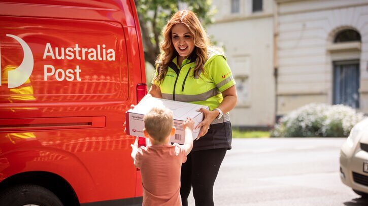 Female Australia Post worker handing parcel to young boy with red Aus Post van in background