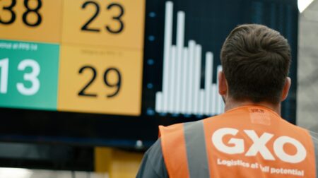 GXO to manage Hisense logistics operations in Spain An employee in an orange GXO high vis jacket stands in front of a board of numbers and graphs.