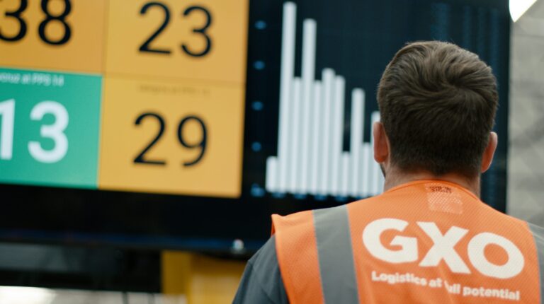 An employee in an orange GXO high vis jacket stands in front of a board of numbers and graphs.