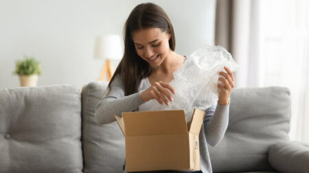 ANALYSIS: From needlessly oversized boxes to labelling, how can parcel packaging be improved? Happy young woman open cardboard box satisfied with purchase online shop order sit on sofa at home, smiling lady customer receive unpack parcel look inside, postal shipping courier service concept