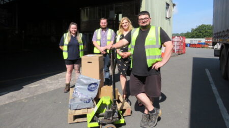 Four logistics workers in high-vis jackets stand in front of a pallet and warehouse.