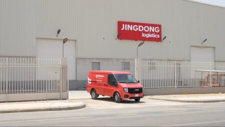 JD Logistics van in front of a grey JD warehouse. The vehicle is exiting the building and turning onto a empty road.
