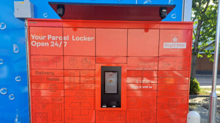 A bank of exterior red parcel lockers with the Royal Mail logo on the top right and a blue wall behind
