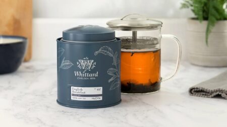 A tin of Whittard tea next to a glass tea strainer with a potted plant on the table behind