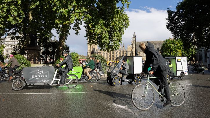 People on a range of bikes - cargo bikes, e-cargo bikes, e-bikes, e-karts – ride along a tree-lined street in London with the Houses of Parliament in the background across the River Thames