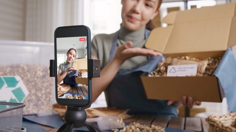 Royal Mail partners with TikTok Shop to streamline deliveries and support UK small businesses A woman performs an unboxing for the camera in an apartment during the day, with a smartphone on a stand on a table and a window behind her