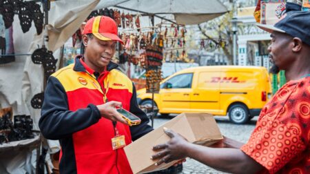 DHL to invest €300m to support African trade growth A DHL worker scans a parcel in a busy street with a yellow, branded DHL van parked behind