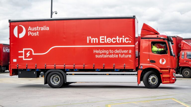A red Australia Post truck with "I'm electric" written down the side sits parked at a depot.