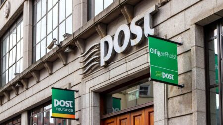 Front of an An Post branch , with green signs hanging out over the street and the post name above the door.