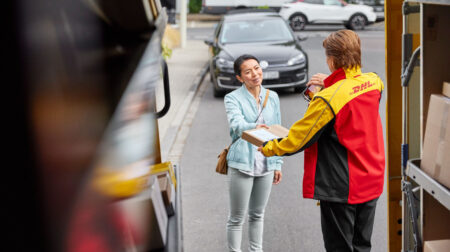 A DHL worker exchanges a parcel with a customer from the back of a delivery van. He is wearing red, yellow and black branded DHL workwear and she is dressed in a light-turquoise jacket and grey pants.