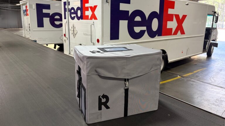 The new reusable box system for B2B shippers from FedEx and Returnity on a conveyor belt at a sorting center. There are FedEx trucks parked in the background.