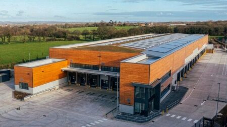 A new DPD regional distribution centers in the southeast of the UK. It has orange brick, grey metal doors and roof and a large forecourt. It is surrounded by fields and trees.