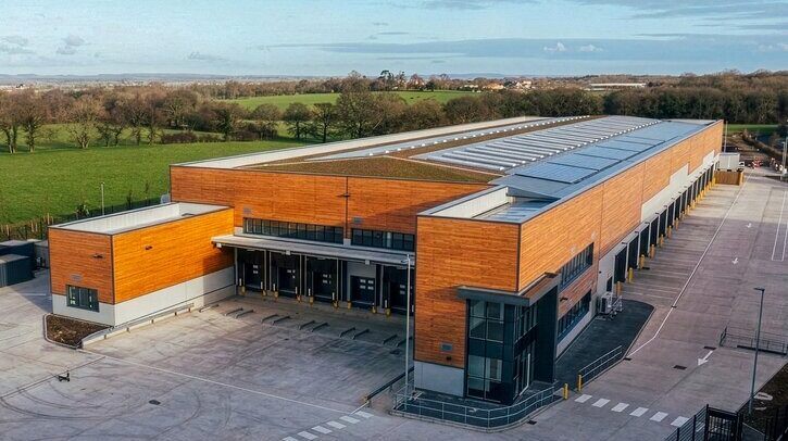 A new DPD regional distribution centers in the southeast of the UK. It has orange brick, grey metal doors and roof and a large forecourt. It is surrounded by fields and trees.