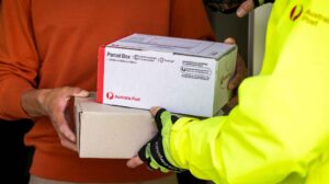 An Australia Post delivery worker wearing a high-vis jacket hands a parcel to a customer wearing an orange jumper and grey trousers. The image shows only their torsos.