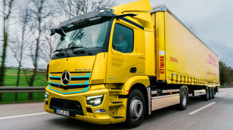 A yellow DHL delivery truck drives along a rural road with grass and grey clouds in the background.