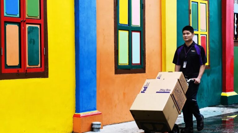 A FedEx delivery worker wheels a stack of boxes past a series of colourful doors and windows.