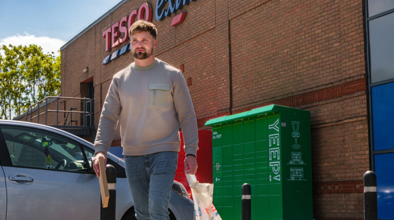 A man walks outside a Tesco store carrying a Tesco carrier bag. A bank of Yeep! parcel lockers is visible behind him
