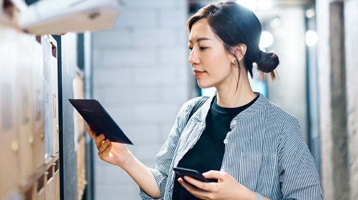 OPINION: Why postal operators are uniquely positioned to own trusted digital communication infrastructure A woman holds up a tablet and a smartphone in front of a large bank of lockers.