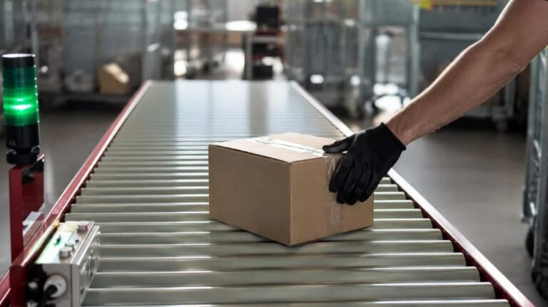 A postal worker wearing black gloves handles a parcel on a conveyor belt in a sorting facility.